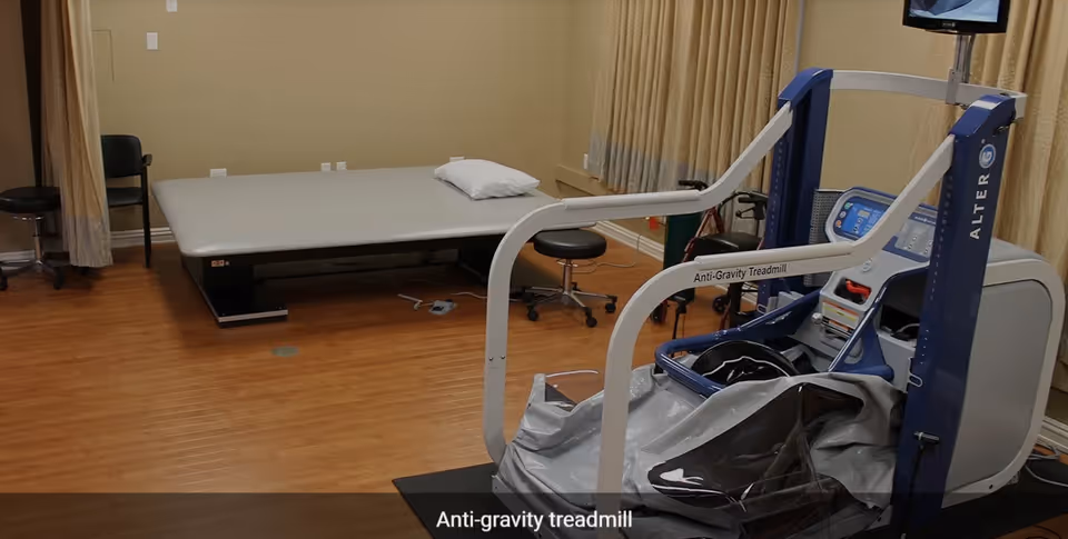 A room with wooden flooring containing an anti-gravity treadmill machine labeled 'ALTER G' and a large padded table with a pillow on it. There are a couple of stools and chairs around the room, and beige curtains cover the windows.