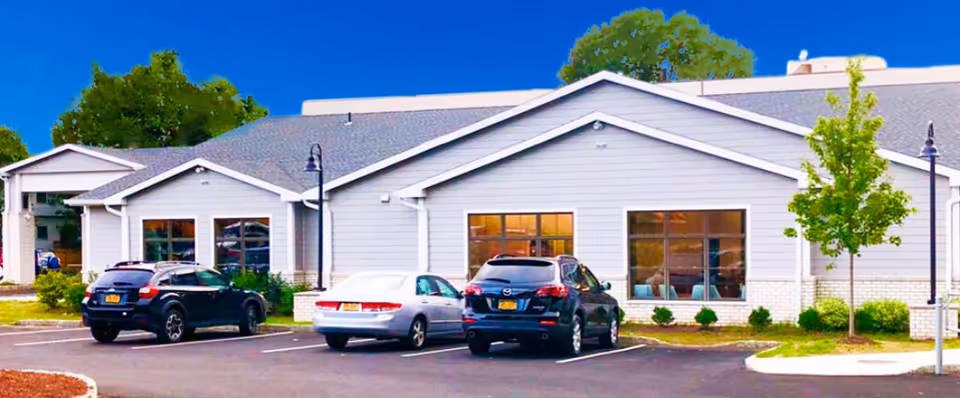 Exterior view of a single-story building with light gray siding and a gabled roof, identified as Tarrytown Rehabilitation and Nursing Center. There are three cars parked in front of the building in a paved parking lot, with small landscaped areas including bushes and a young tree. The sky is clear and blue.