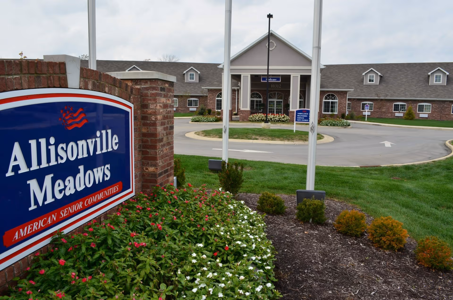 Entrance to Allisonville Meadows senior living community showing a brick sign with the community name and a large building in the background with a circular driveway and landscaped greenery.