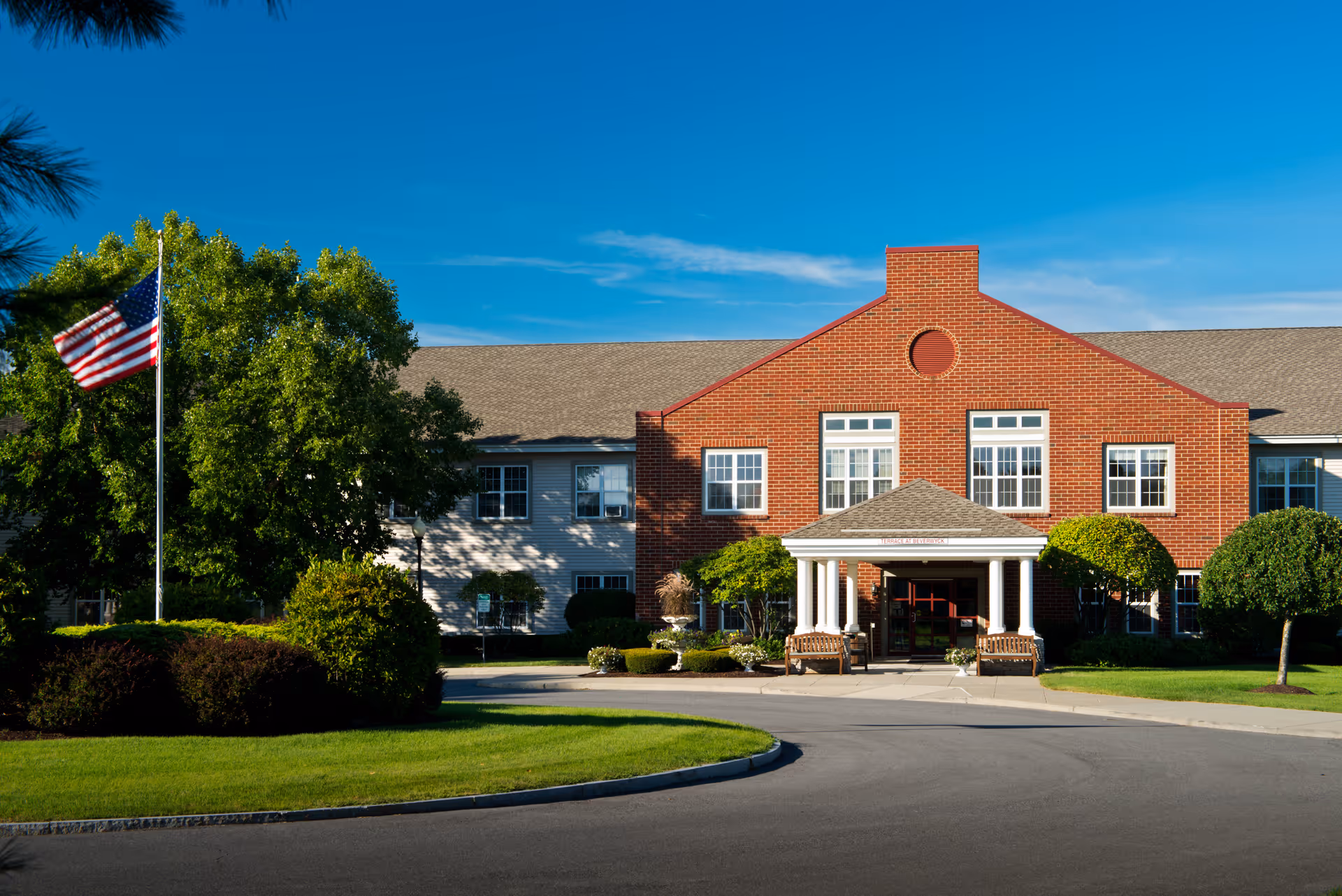 Front exterior view of a senior living facility named Beverwyck with a brick facade, white columns at the entrance, well-maintained landscaping, and an American flag on a flagpole to the left.
