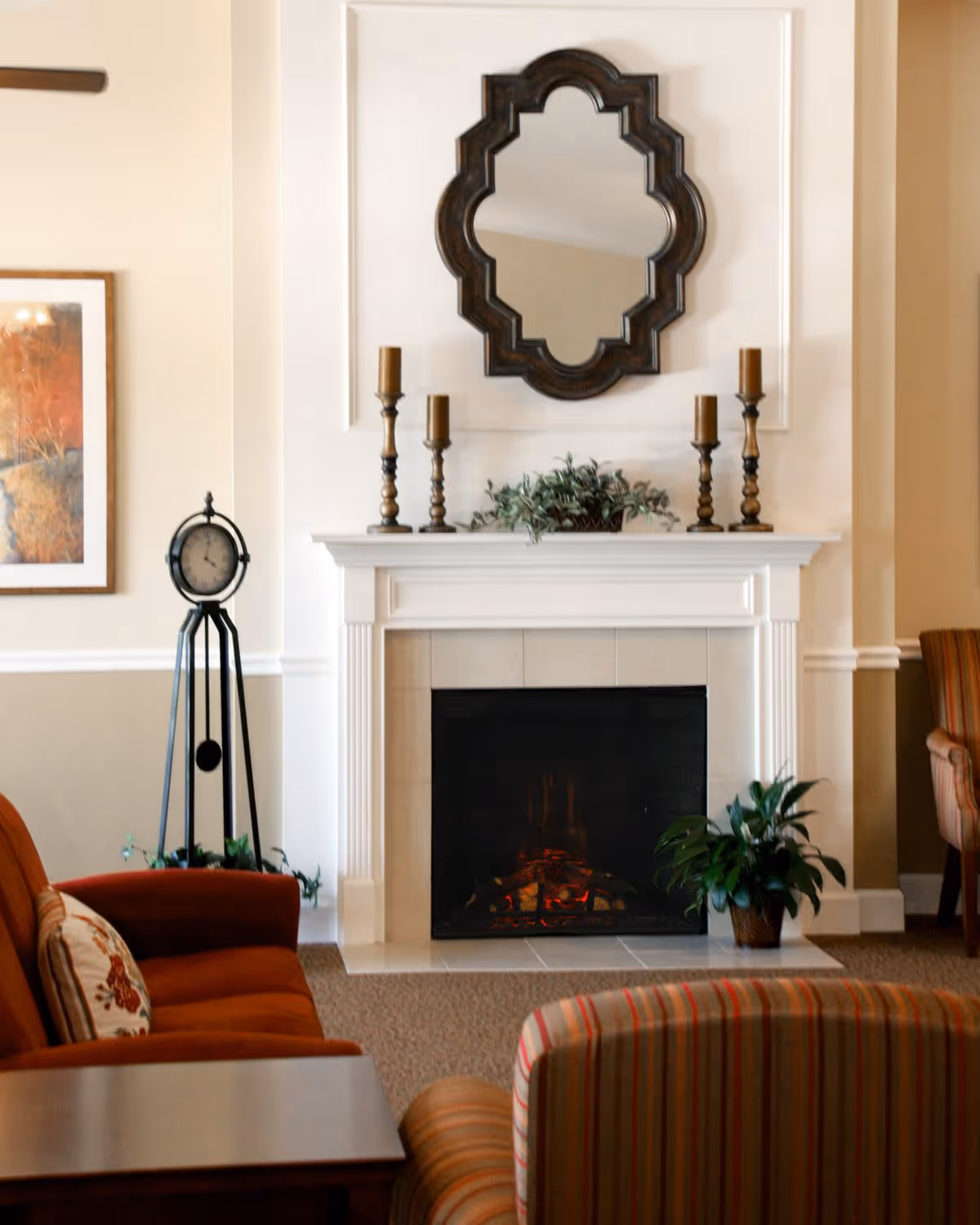 A cozy living room area with a white fireplace featuring a decorative mirror above it, five candle holders, and a green plant on the mantel. There is a standing clock to the left, a framed painting on the wall, and comfortable upholstered chairs and a sofa in warm tones around a wooden table.