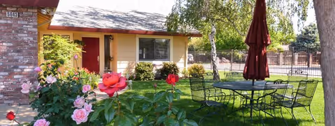 Outdoor garden area of a senior living facility with blooming pink and red roses in the foreground, a round metal table with six chairs and a closed red umbrella on a grassy lawn, and a single-story building with a red door and windows in the background.