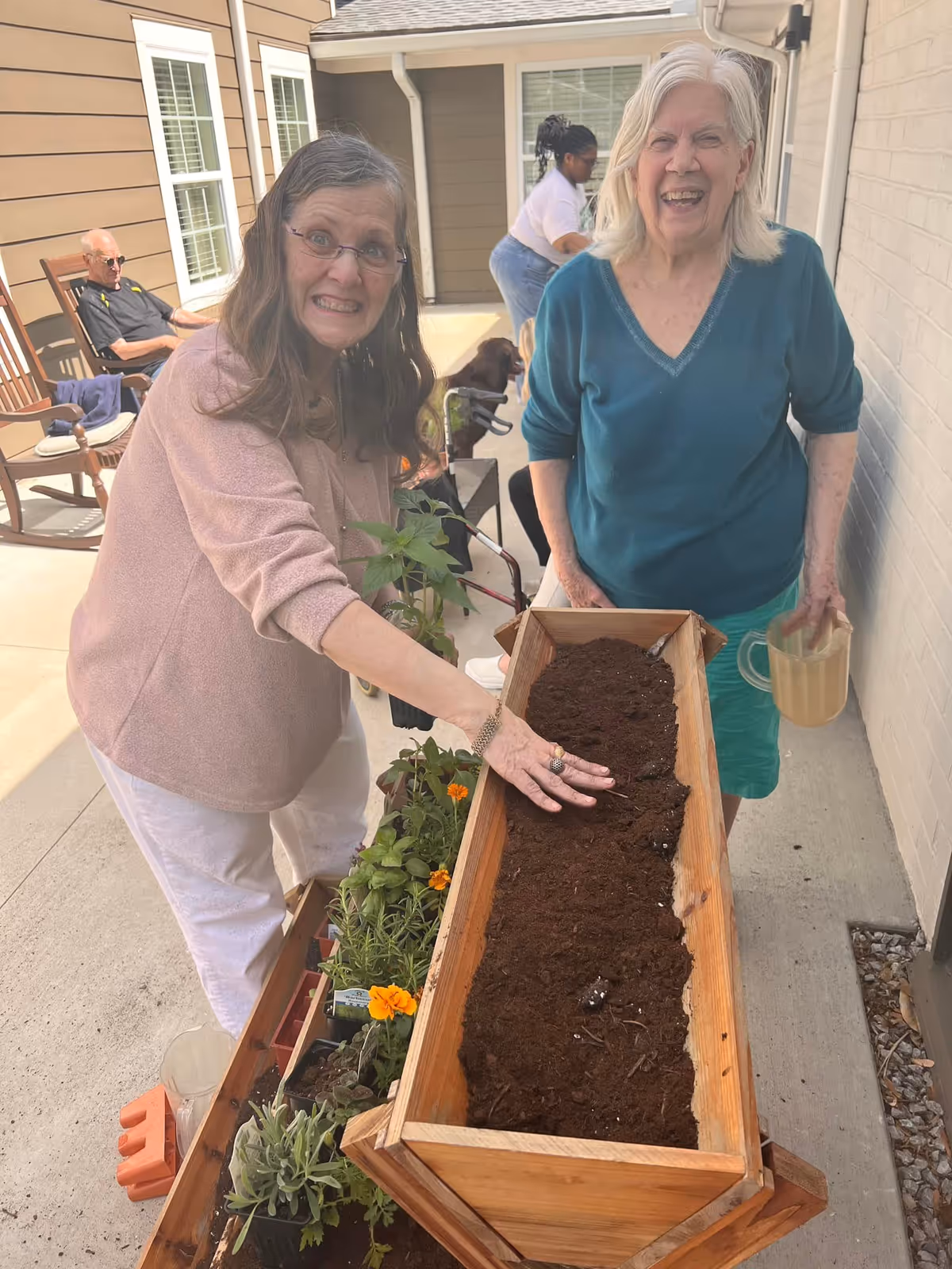 Two elderly women smiling and engaging in gardening activity on a patio. One woman is touching soil in a wooden planter box filled with soil, while the other holds a watering can. In the background, a man is sitting on a rocking chair and another person is tending to a dog.