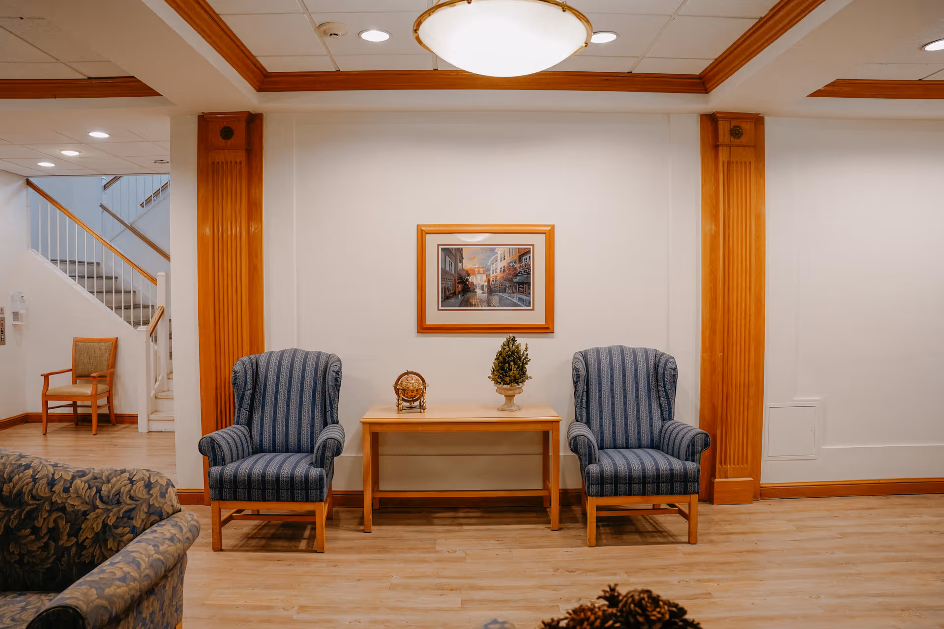 A cozy seating area in a senior living facility with two blue striped armchairs placed on either side of a wooden table. On the table, there is a small decorative globe and a potted plant. Above the table hangs a framed picture of a street scene. The room has wooden flooring, white walls, and wooden trim accents. In the background, there is a staircase and an additional wooden chair.