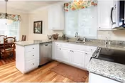 Bright kitchen with white cabinets, granite countertops, stainless steel dishwasher and stove, hardwood floors, a window with floral valance above the sink, and a dining area with wooden chairs and table in the background.