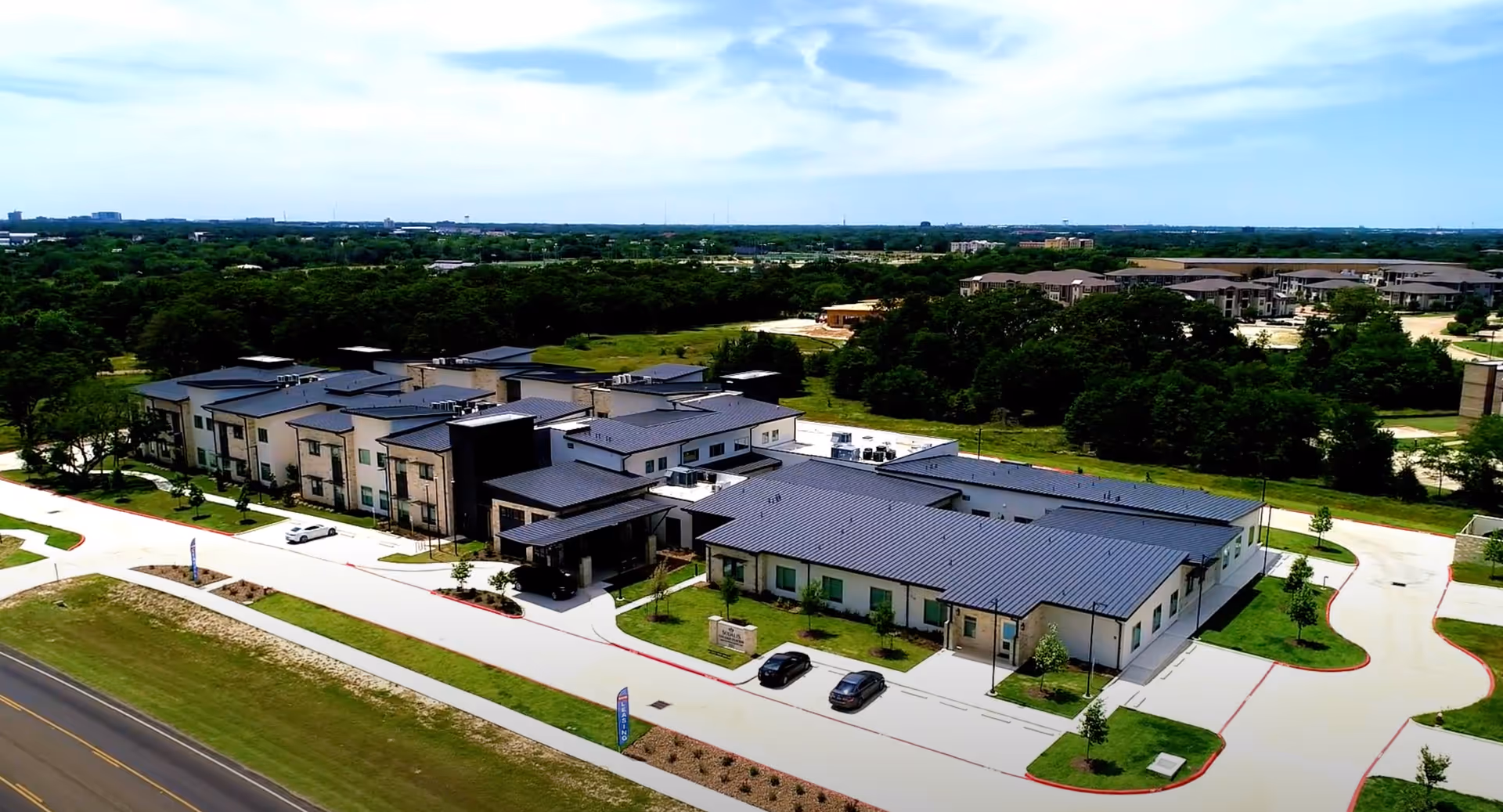 Aerial view of a large, modern senior living facility building with multiple sections and a dark roof, surrounded by green lawns, trees, and parking areas under a partly cloudy sky.