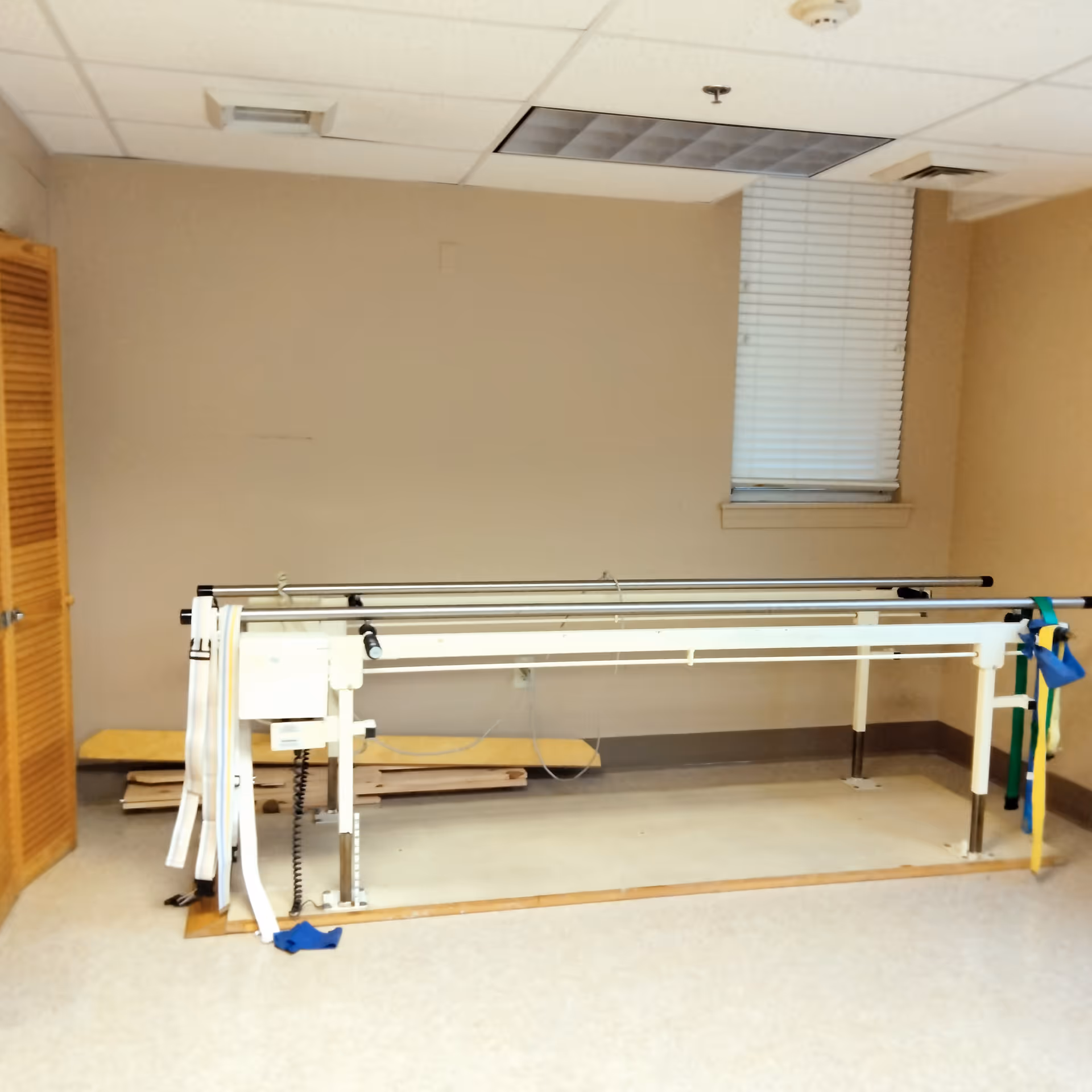 A rehabilitation room with a parallel bar setup used for physical therapy. The room has beige walls, a window with closed blinds, a wooden door on the left, and some therapy straps hanging on the bars.