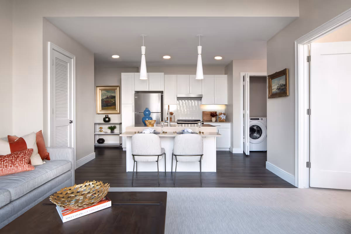A modern open-concept living area and kitchen in a senior living facility. The living room has a gray sofa with red and beige pillows, and a dark wooden coffee table with a decorative gold bowl and books. The kitchen features white cabinetry, a stainless steel refrigerator, a stove with a hood, and a central island with two light gray chairs. Pendant lights hang above the island. To the right, a laundry area with a washing machine is visible through an open door. The walls are painted light beige, and the flooring is dark wood with a light-colored rug in the living area.