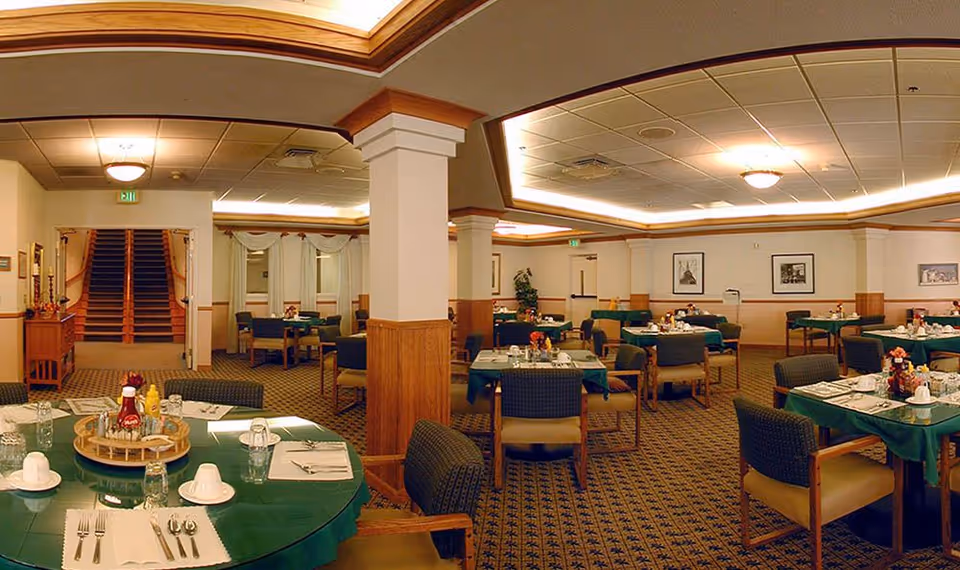 A spacious dining room in a senior living facility with multiple round tables covered with green tablecloths. Each table is set with white plates, cups, silverware, and condiments. The room features carpeted floors, wooden paneling on the walls, framed pictures, and a ceiling with recessed lighting. There is a staircase visible in the background.