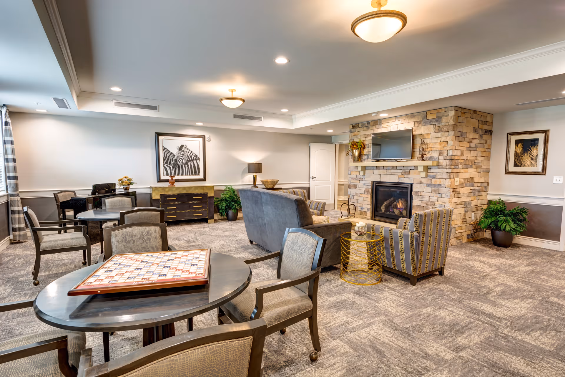 A cozy senior living common area with a stone fireplace, a mounted TV above it, and comfortable seating including a sofa and two armchairs. There are multiple tables with chairs around them, one table has a Scrabble board game on it. The room is well-lit with ceiling lights and has framed artwork on the walls and potted plants for decoration.