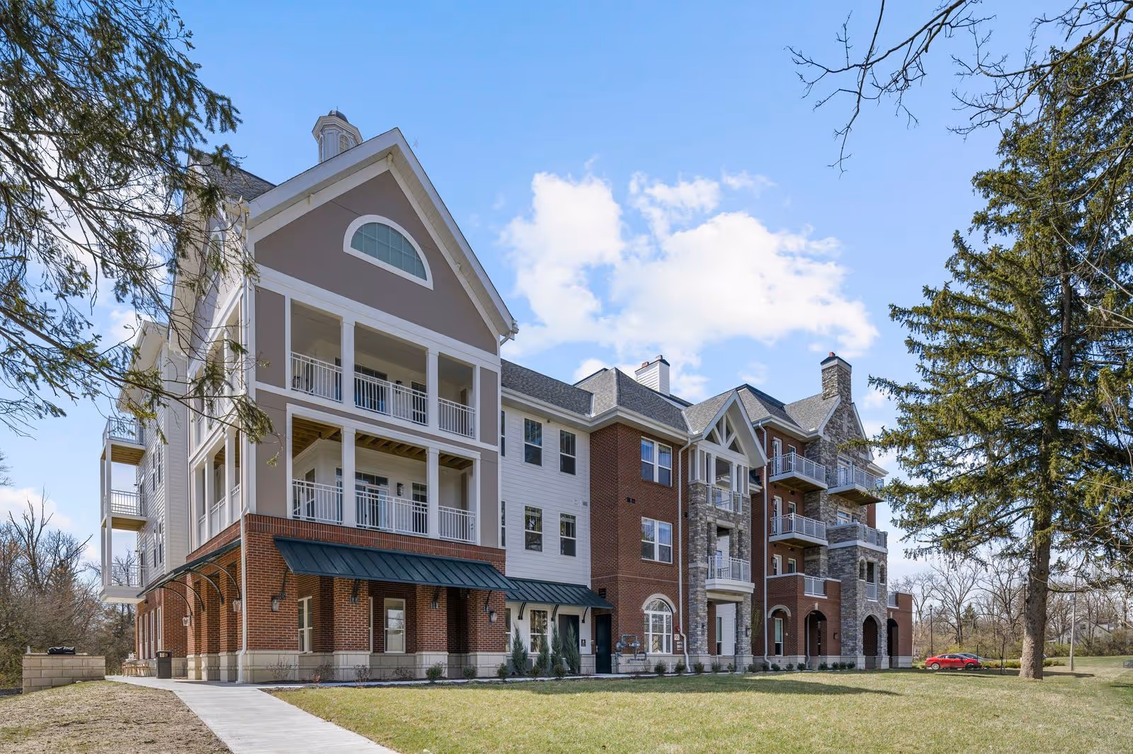Exterior view of a multi-story senior living facility building with a combination of brick, stone, and siding. The building features balconies, large windows, and a green awning over the entrance. There are trees and a grassy lawn in the foreground under a blue sky with some clouds.