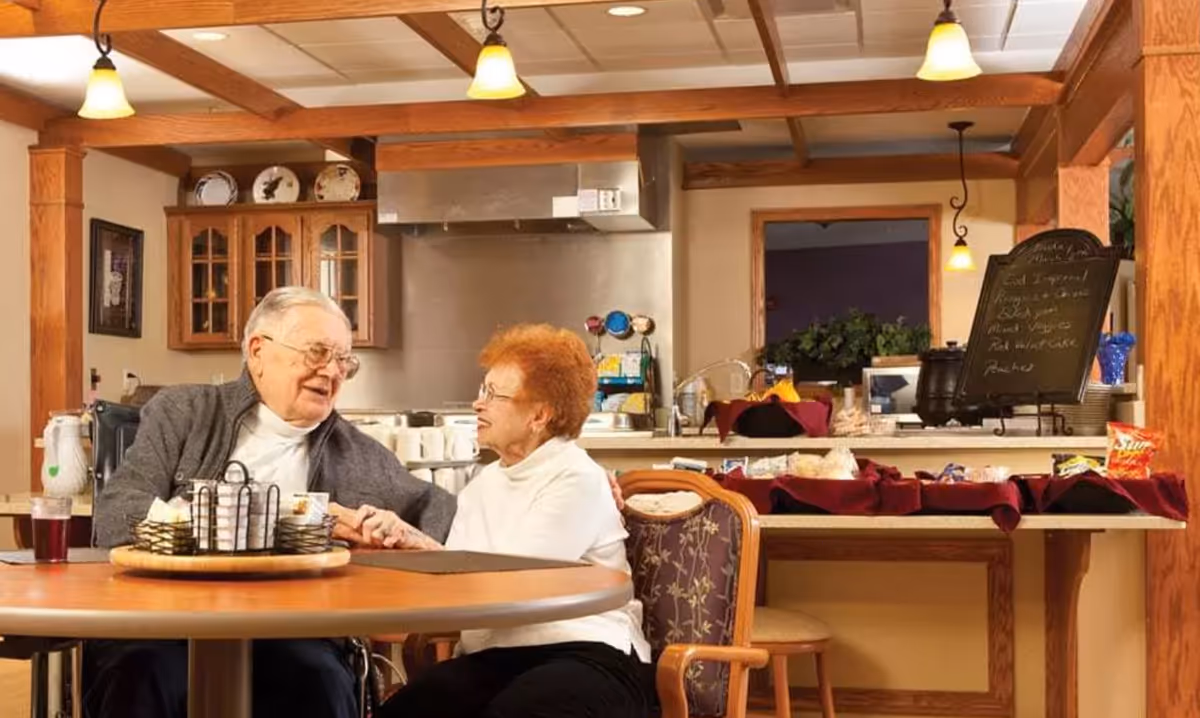 An elderly man and woman sitting at a round wooden table in a warm, well-lit dining area. The man is in a wheelchair and they are holding hands and smiling at each other. Behind them is a kitchen counter with various food items and a small chalkboard menu. The room features wooden beams on the ceiling and wooden cabinetry.
