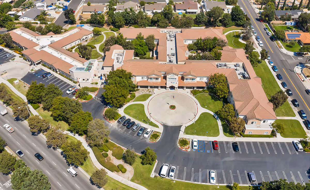 Aerial view of Bayshire Yorba Linda senior living facility showing a large building with a circular driveway entrance, surrounded by green lawns, trees, parking lots, and nearby streets with cars.