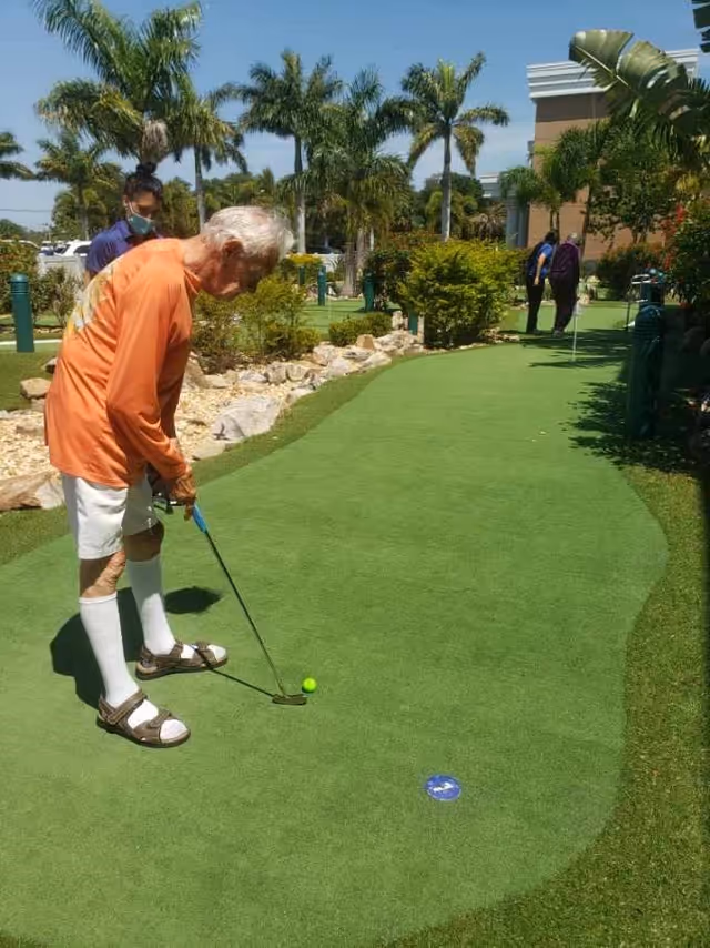 An elderly man wearing an orange long-sleeve shirt, white shorts, knee-high white socks, and sandals is playing mini golf on a green putting surface outdoors. There are palm trees, bushes, and rocks surrounding the area. In the background, two other people are visible near the putting green, and a building is partially visible on the right side.