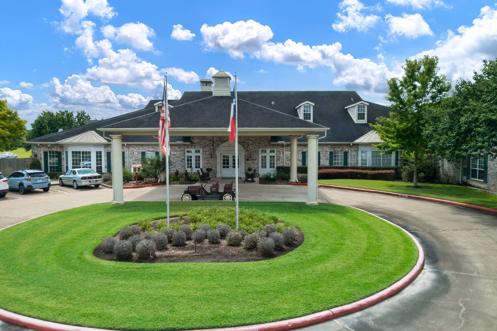 Front exterior view of Trinity Oaks of Pearland facility showing a circular driveway with a landscaped island in the center, two flagpoles with American and Texas flags, a covered entrance supported by columns, and several parked cars. The building has a brick facade, multiple windows, and a dark shingled roof under a partly cloudy blue sky.