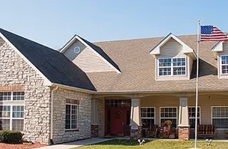 Exterior view of a senior living facility building with stone and siding facade, multiple windows, a front entrance with a red door, and an American flag on a flagpole in front.