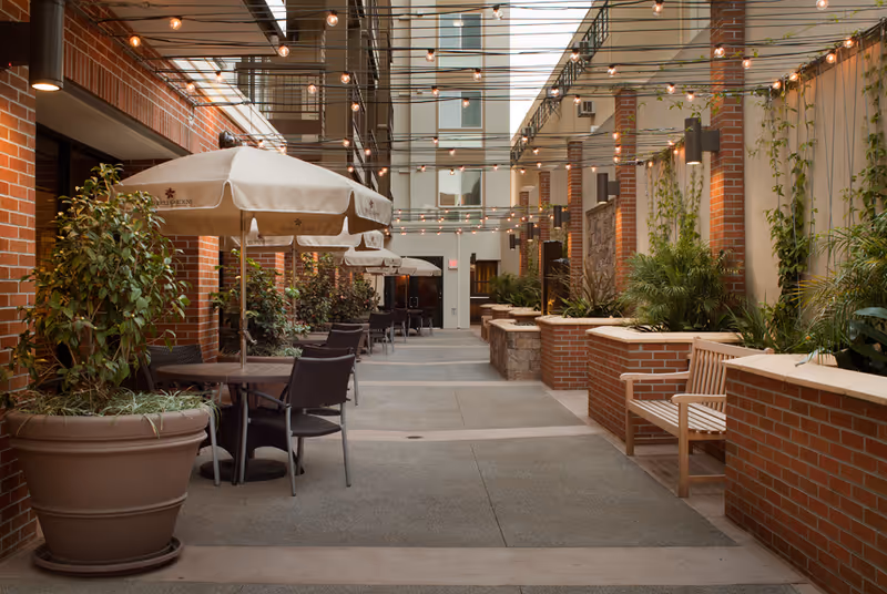 Outdoor courtyard area with brick walls and planters, string lights overhead, tables with umbrellas and chairs along one side, and wooden benches along the other side, surrounded by greenery.