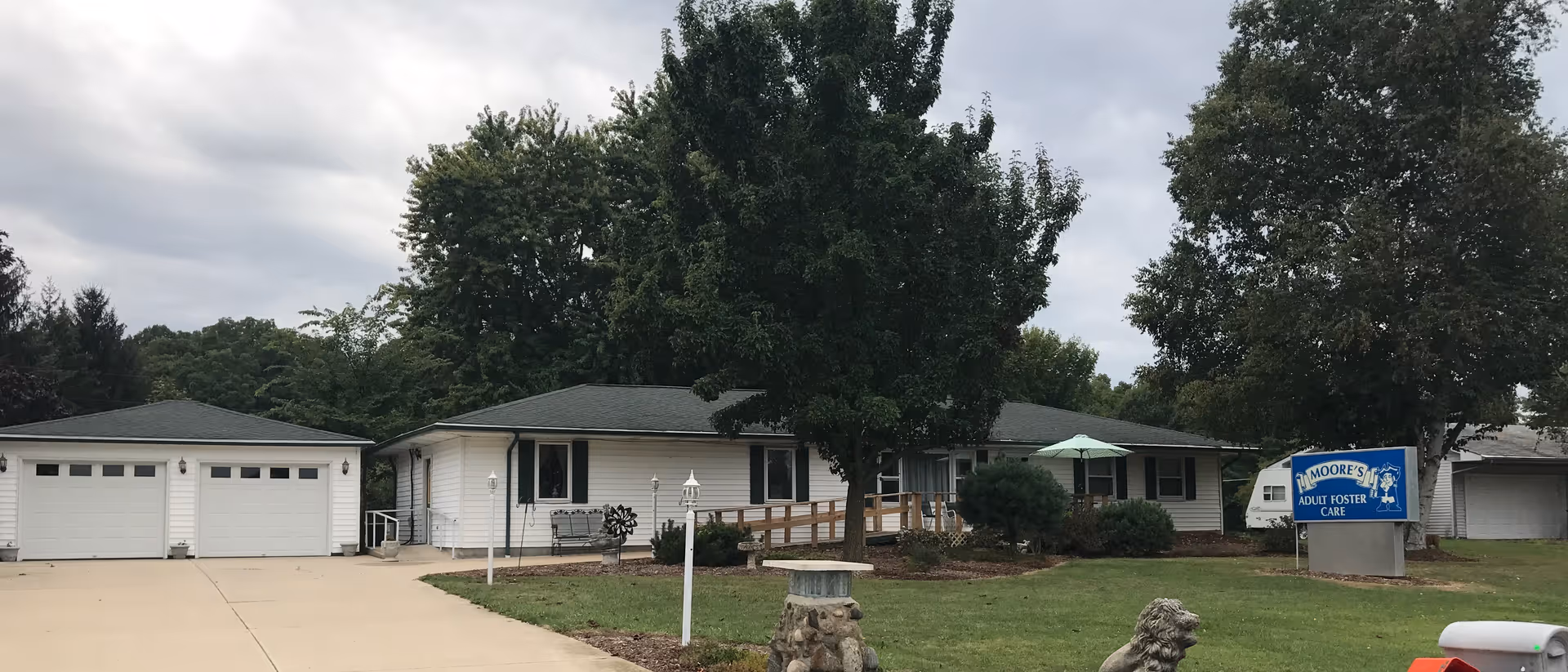 Exterior view of Moore's Adult Foster Care facility showing a single-story white building with a dark roof, surrounded by trees and greenery. There is a driveway leading to a two-car garage on the left, a lawn with landscaping in front, and a blue sign on the right that reads 'Moore's Adult Foster Care'.