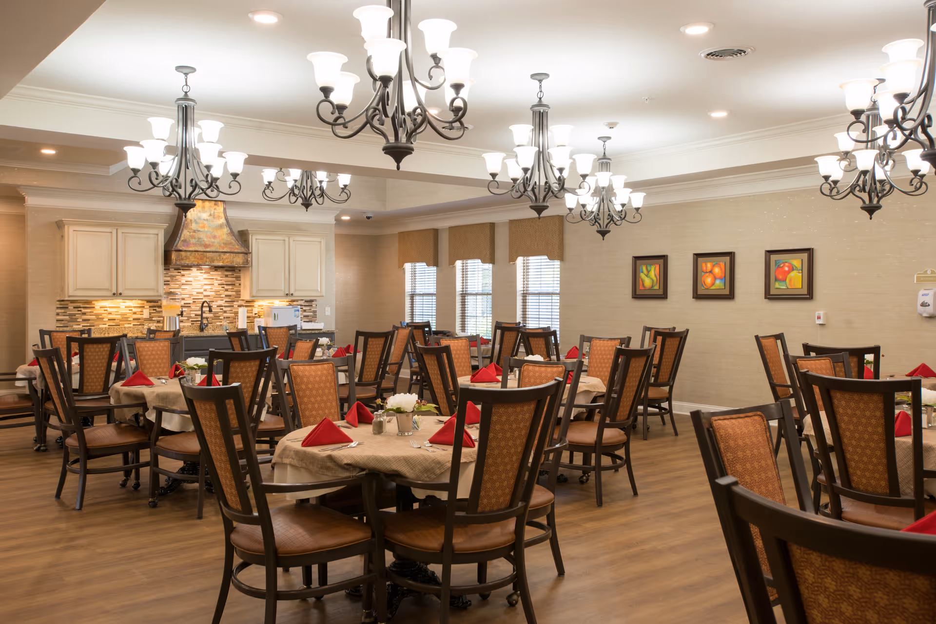A well-lit dining room with multiple round tables covered with beige tablecloths, each set with red folded napkins and small floral centerpieces. The room features wooden chairs with patterned upholstery, chandeliers hanging from the ceiling, and a kitchen area with cabinets and a tiled backsplash in the background. Three framed fruit paintings hang on the wall.