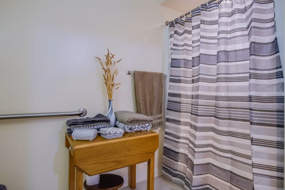 A bathroom corner featuring a wooden table with folded towels and a vase with dried plants on top. A towel hangs on a wall-mounted towel rack next to a striped shower curtain. A metal grab bar is attached to the wall.