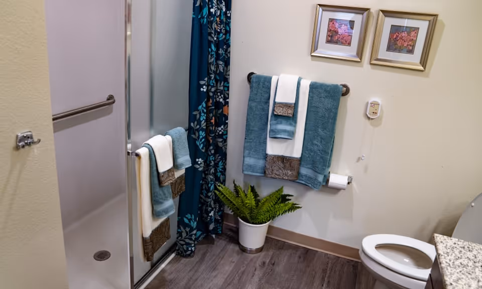 A bathroom with a walk-in shower featuring a glass door and a floral shower curtain. There are teal and white towels hanging on towel racks, a potted green plant on the floor, two framed floral pictures on the wall, a toilet, and a granite countertop partially visible.