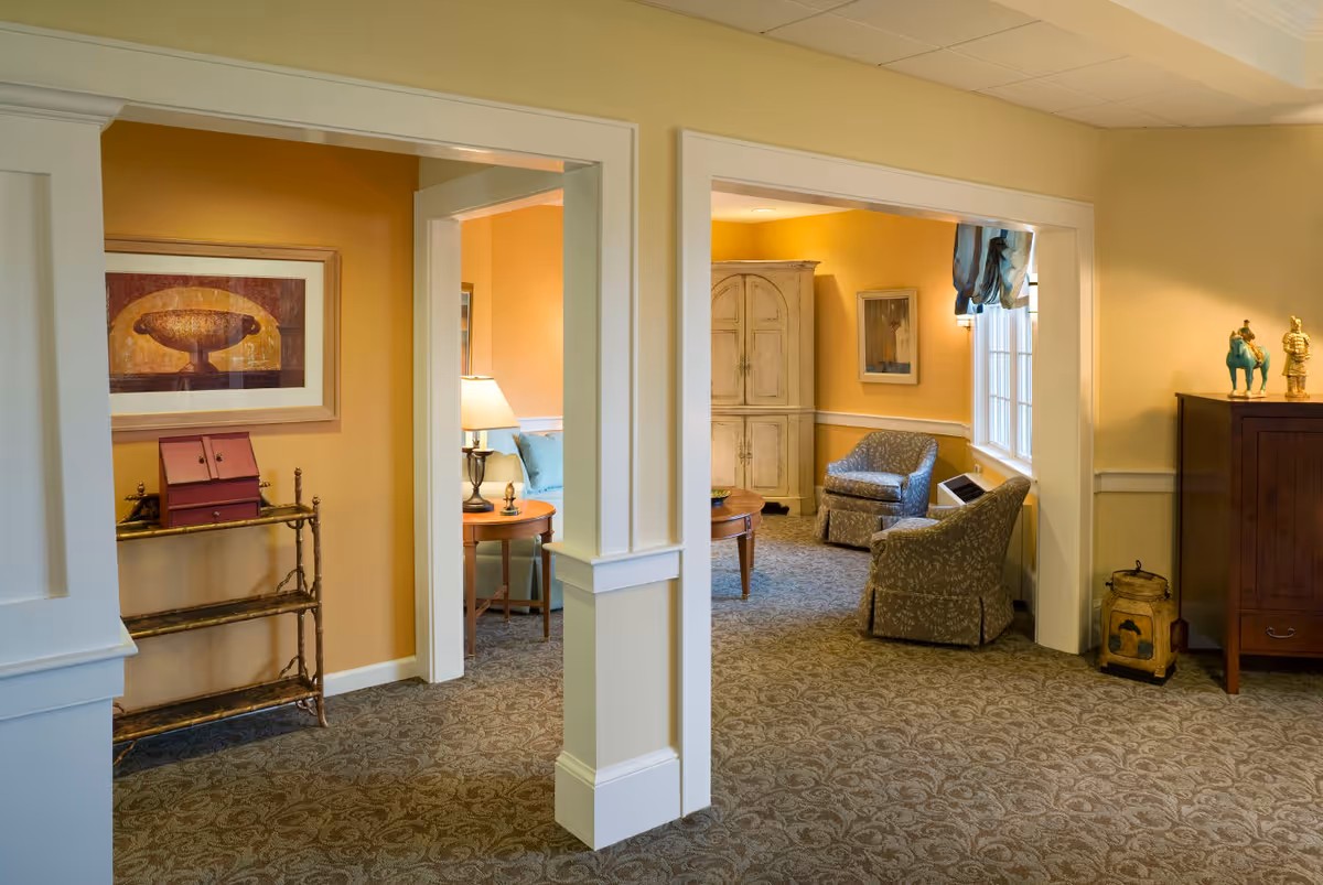 Interior view of a senior living facility showing a cozy sitting area with two armchairs, a wooden cabinet, and a small table with a lamp. The walls are painted yellow and decorated with framed artwork. The carpet has a patterned design, and there is a decorative shelf with items on it.