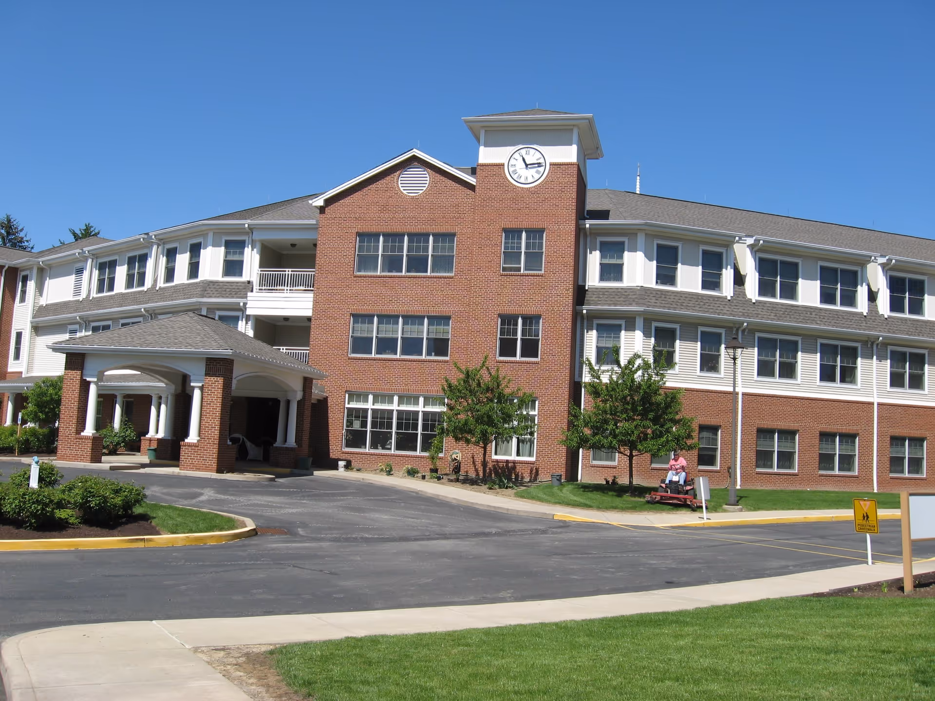 Exterior view of a multi-story brick and white siding building with a clock tower, a covered entrance, and a person riding a lawn mower on the grass near the building under a clear blue sky.