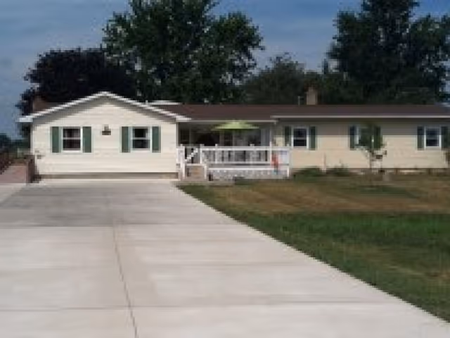 Single-story residential building with a wide concrete driveway leading up to the front porch. The house has light-colored siding with green shutters on the windows and a brown roof. There are trees in the background and a grassy lawn on either side of the driveway.