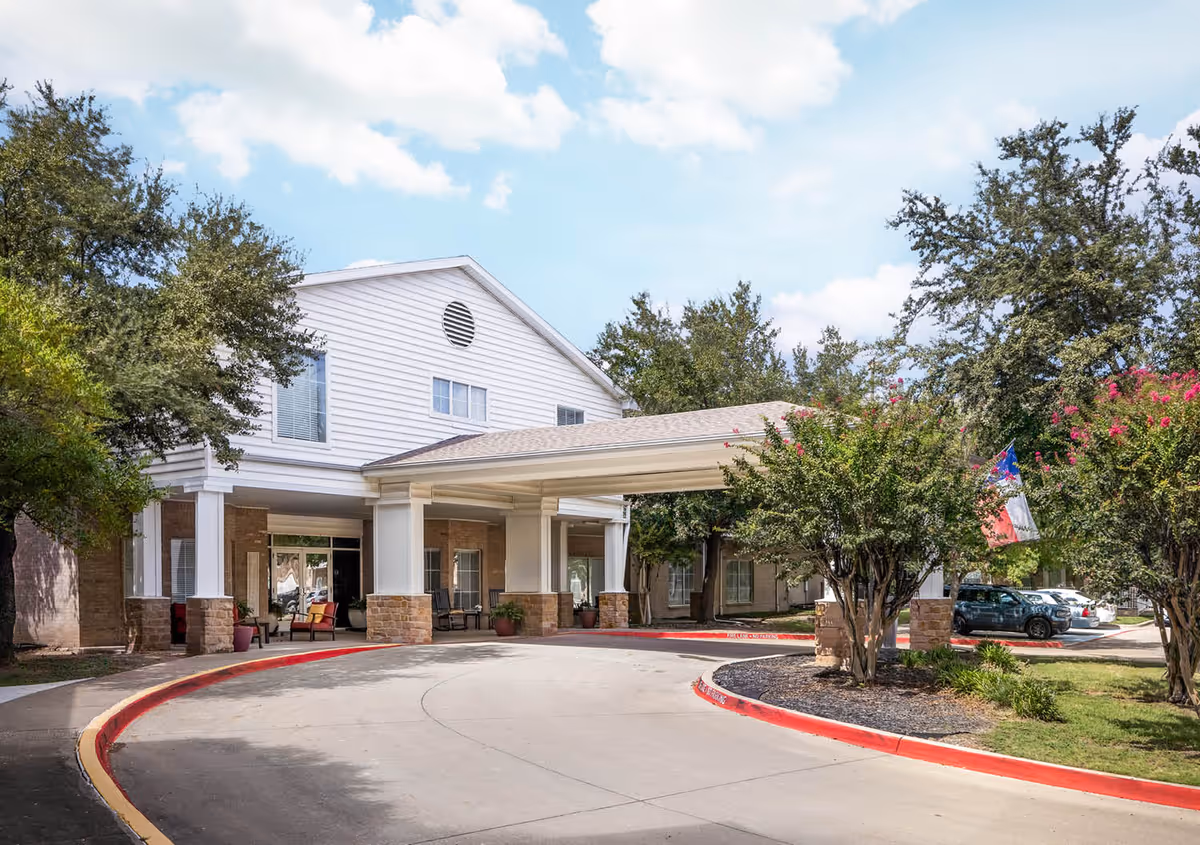 Exterior view of The Wellington at North Richland Hills senior living facility showing the entrance with a covered driveway, surrounded by trees and parked cars under a partly cloudy sky.