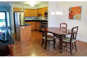 Interior view of a dining area and kitchen in a senior living facility. The dining area features a wooden table with four chairs, and a hanging light fixture above. The kitchen has wooden cabinets, a stainless steel refrigerator, stove, and a black countertop. The floor is wooden, and there is a colorful painting on the wall near the dining table.