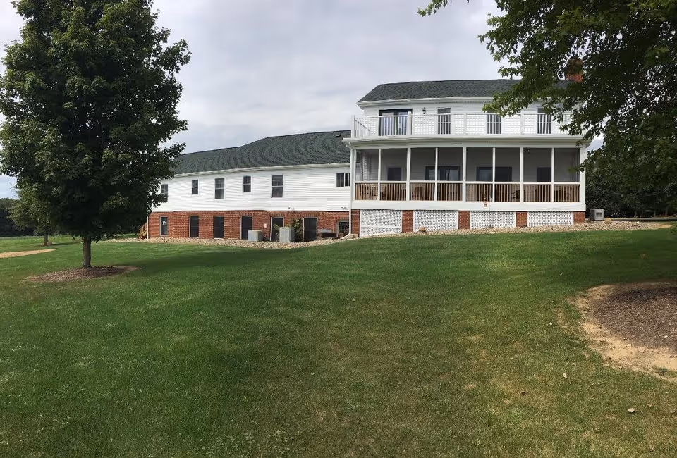 Exterior view of a two-story assisted living facility building with white siding and a green roof, surrounded by a well-maintained grassy lawn and trees under a cloudy sky.