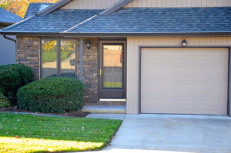 Exterior view of a residential unit at Creekside at Elfindale Independent Living showing a front door with a glass storm door, a window with blinds, a garage door, and neatly trimmed bushes beside a green lawn.
