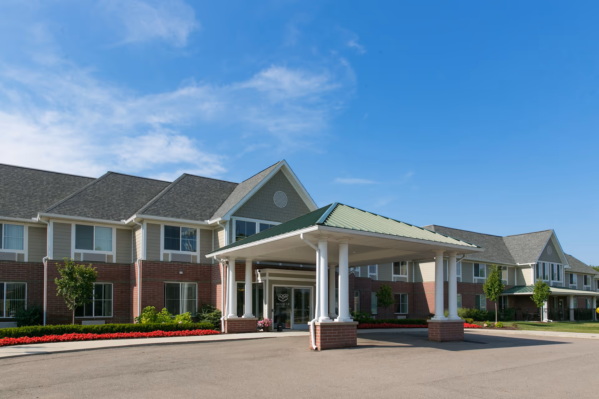 Exterior view of Belmar Oakland senior living facility showing a two-story building with a covered entrance, brick and siding facade, landscaped with small trees and red flowers under a clear blue sky.