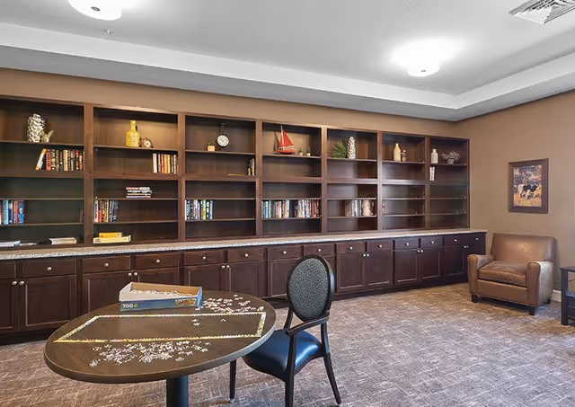 A cozy room with a large wooden bookshelf filled with books and decorative items along the back wall. In front, there is a round table with a partially completed jigsaw puzzle and a single chair. To the right, there is a brown leather armchair and a framed picture on the wall. The room has carpeted flooring and soft lighting from ceiling fixtures.