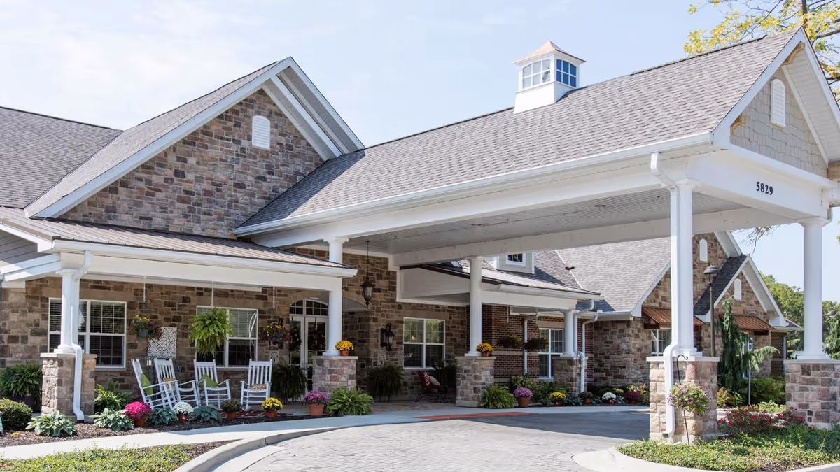 Exterior view of a senior living facility with a covered entrance supported by white columns. The building features stone and brick walls, multiple windows, and a gray shingled roof. There are several white rocking chairs on the porch, surrounded by potted plants and colorful flowers.