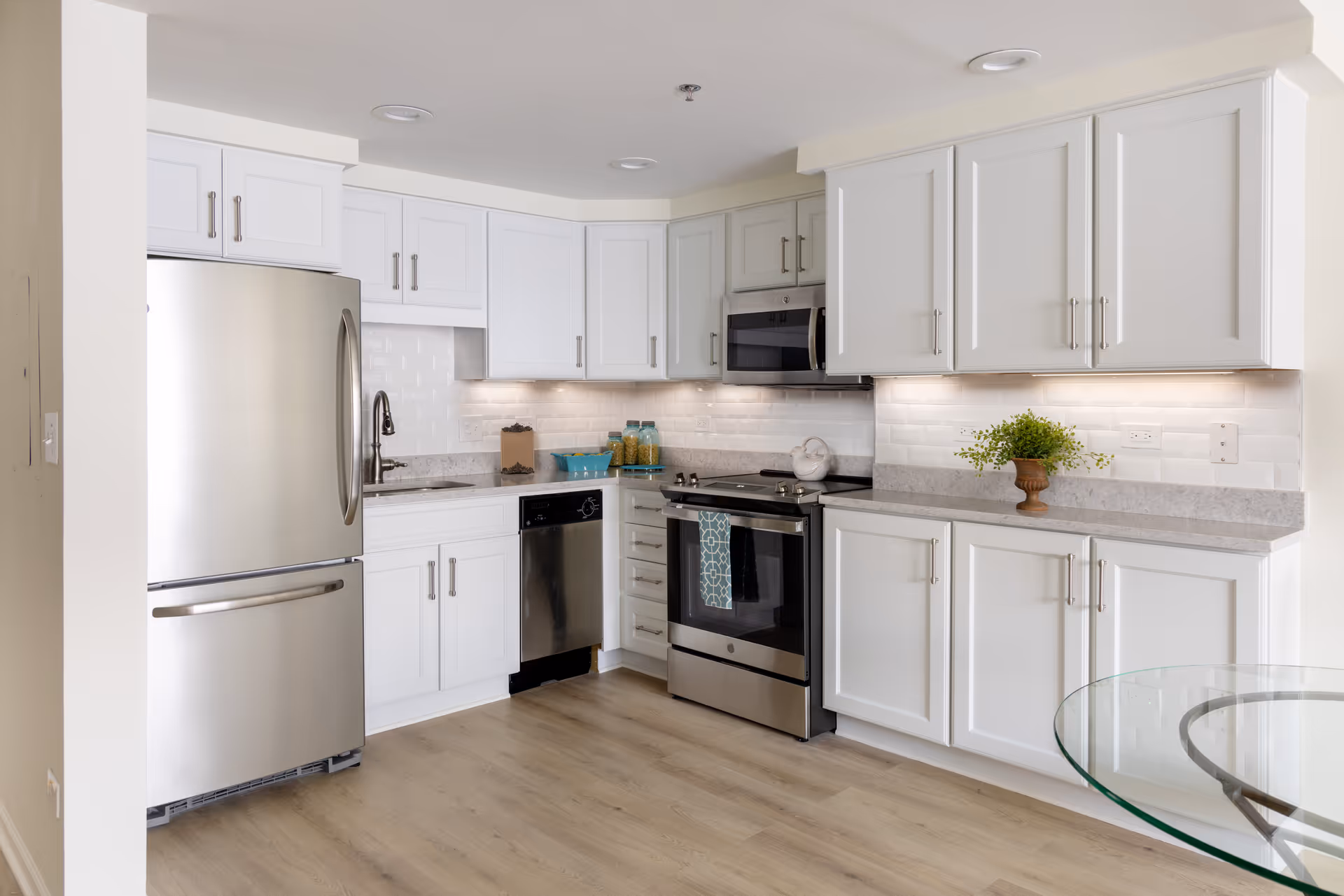 Modern kitchen with white cabinets, stainless steel refrigerator, dishwasher, oven, and microwave. Light wood flooring and a glass-top table partially visible on the right. Under-cabinet lighting illuminates the white subway tile backsplash.