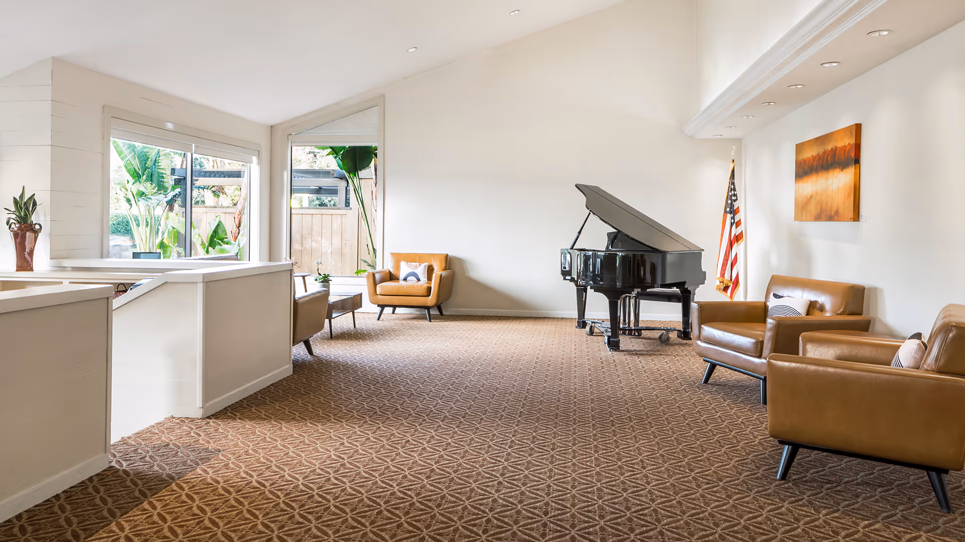 A spacious, well-lit living room area with a grand piano, several brown leather armchairs, a small coffee table, an American flag, and a large window showing greenery outside. The room has patterned carpet and light-colored walls with a piece of artwork hanging.