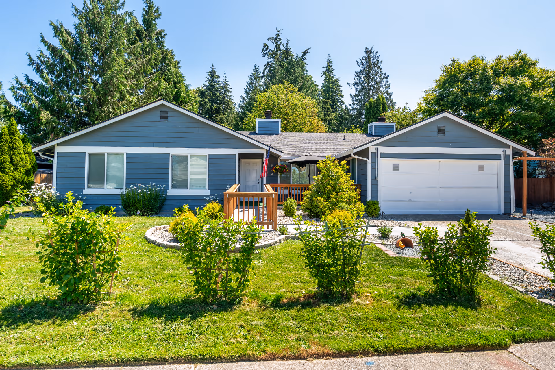 Front exterior view of a single-story blue house with white trim, a two-car garage, a small porch with an American flag, and a well-maintained green lawn with bushes and trees in the background under a clear blue sky.