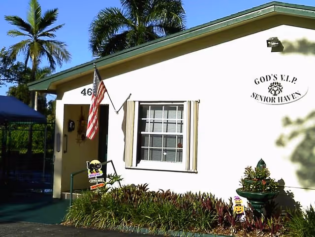 Exterior view of a single-story building with a white facade and green roof trim. There is an American flag mounted near the entrance, a window with beige shutters, and landscaping with green plants and bushes in front. The building has a sign that reads 'GOD'S V.I.P SENIOR HAVEN'.