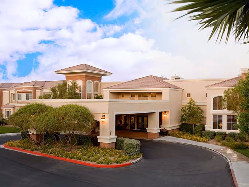 Exterior view of a senior living facility named Atria Seville, showing a beige building with a covered entrance, landscaped greenery, and a curved driveway under a partly cloudy blue sky.