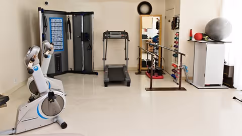 A small exercise room with a stationary bike, treadmill, weight machine, parallel bars for walking support, and shelves holding exercise balls and weights.