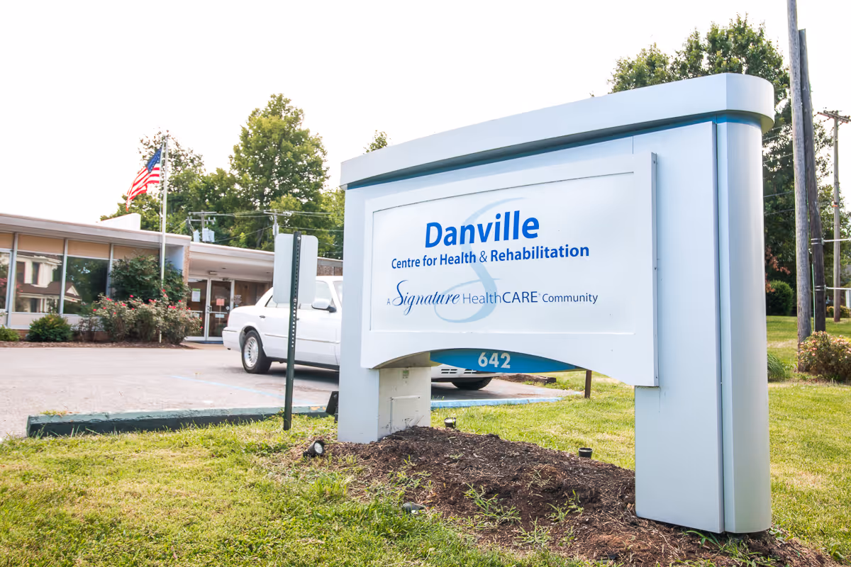 Outdoor view of the Danville Centre for Health and Rehabilitation sign with the building and an American flag in the background. A white car is parked near the entrance, and there is grass and some bushes around the area.