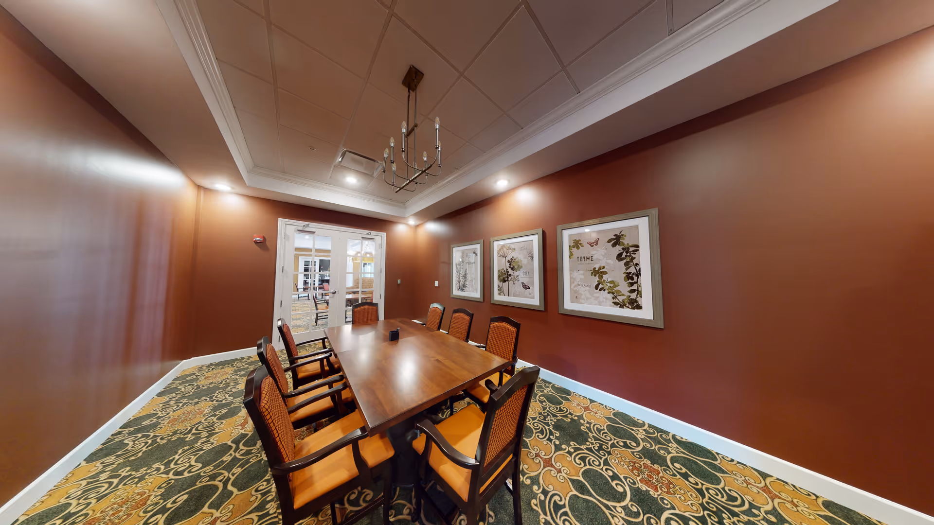 A formal dining room with a long wooden table surrounded by eight cushioned chairs with orange upholstery. The walls are painted a warm brown color and decorated with three framed botanical prints. The ceiling features recessed lighting and a chandelier. The floor is covered with a patterned carpet in green, orange, and beige tones. Double glass doors lead to another room in the background.