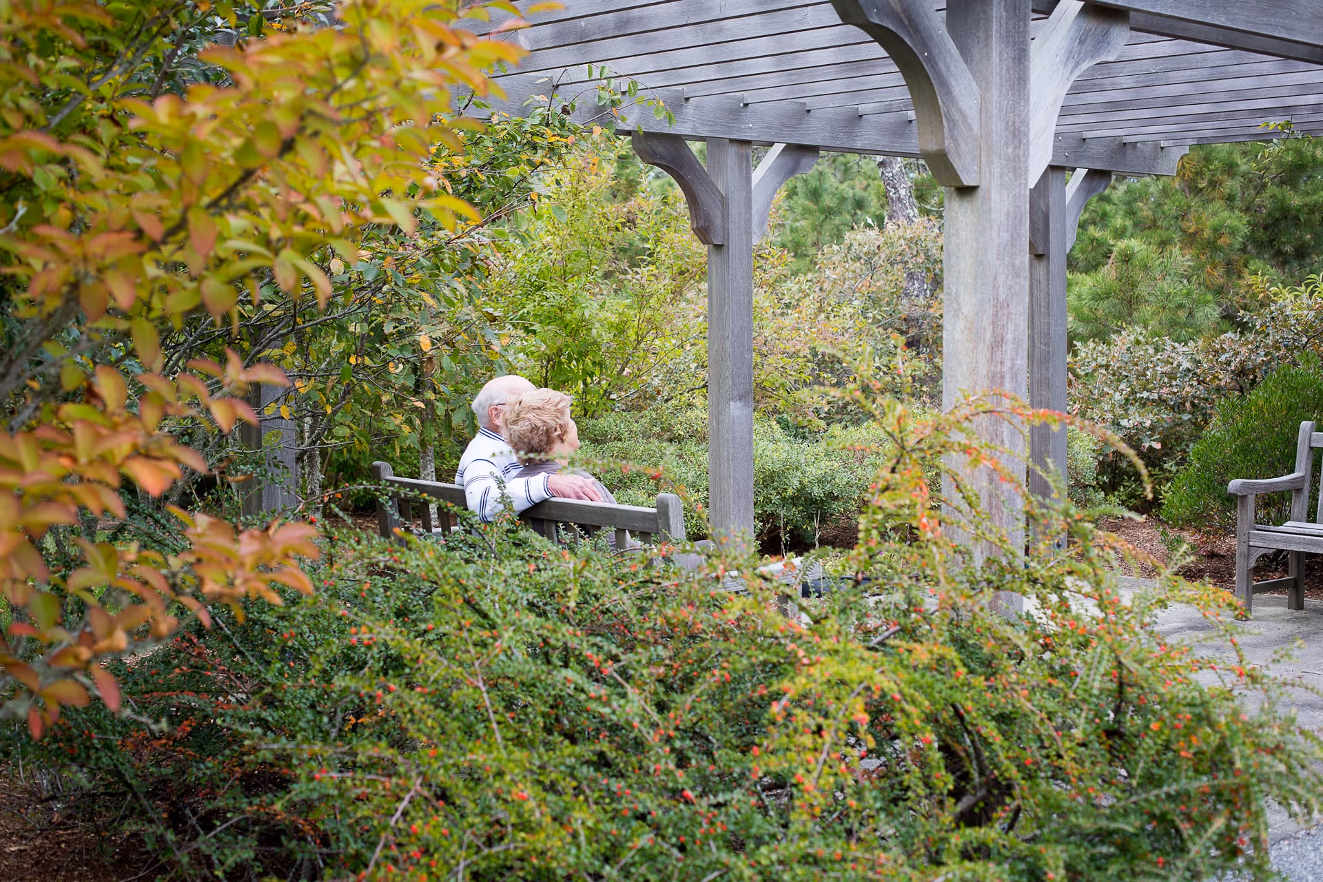 An elderly couple sitting closely together on a wooden bench under a wooden pergola in a lush garden area surrounded by green shrubs and trees.