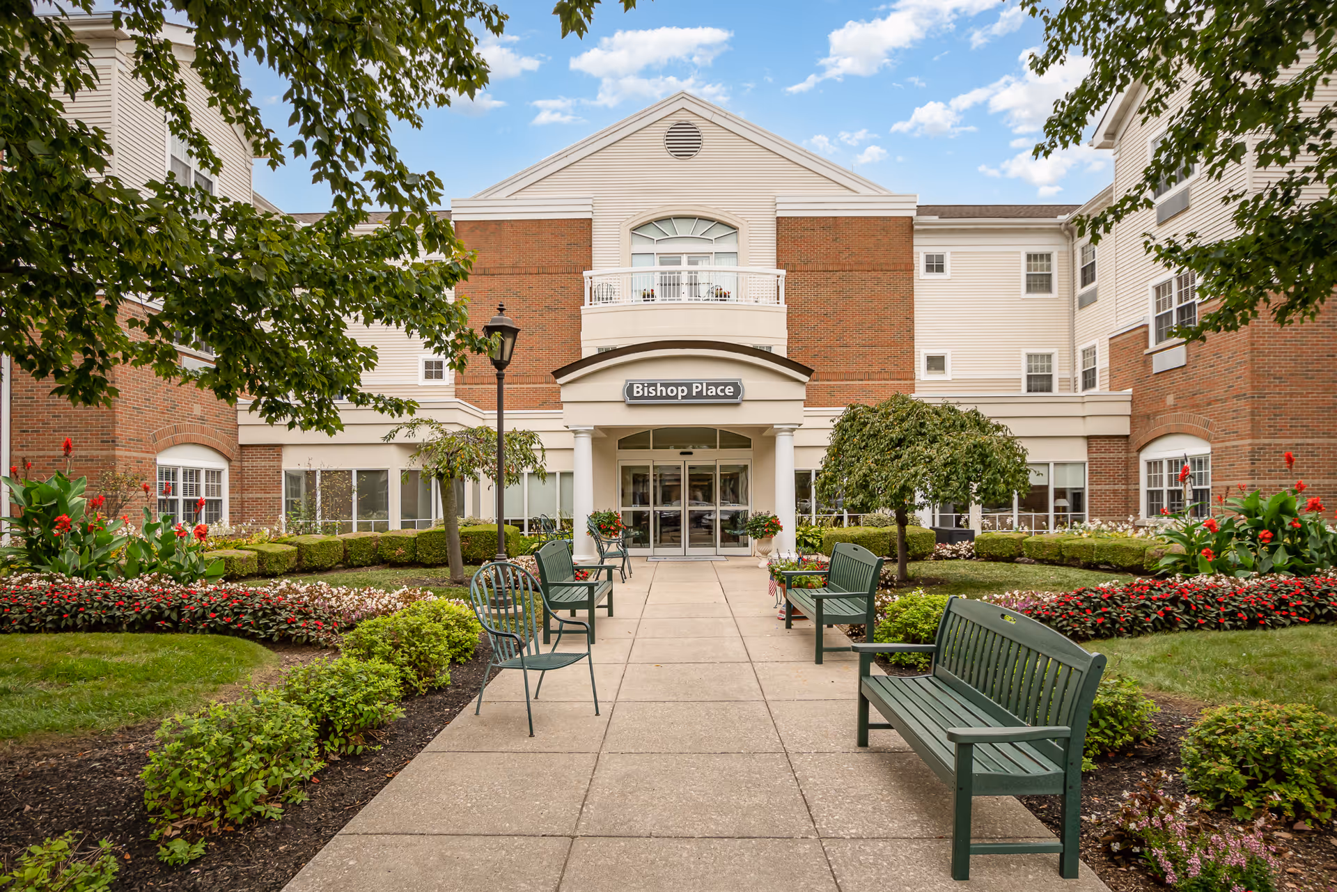Front exterior view of a senior living facility named Bishop Place, featuring a brick and beige building with a central entrance, surrounded by well-maintained gardens, green benches, and trees under a partly cloudy sky.