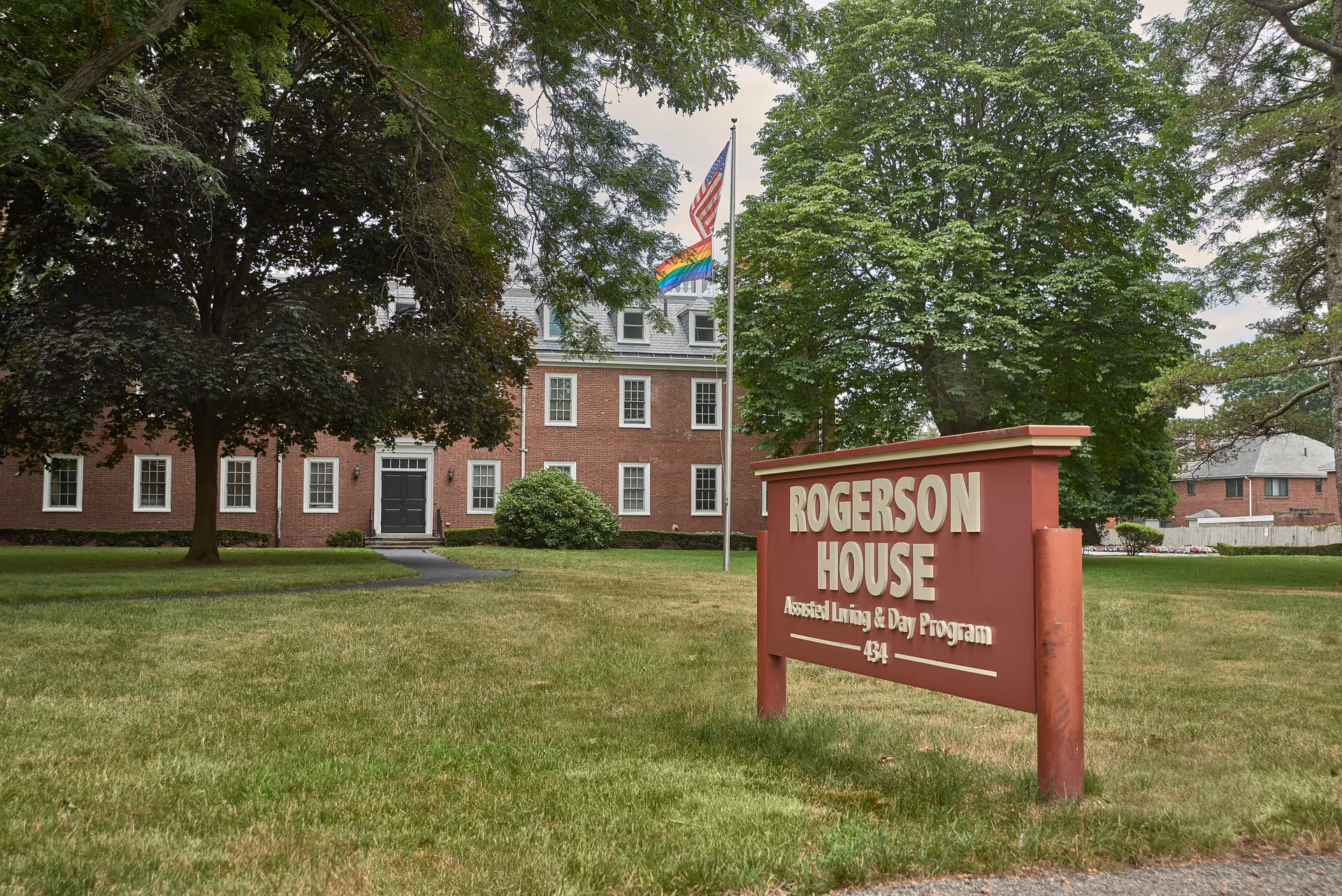 Exterior view of Rogerson House Memory Care Assisted Living & Day Program, showing a large brick building with multiple windows, a black front door, and a well-maintained lawn with trees. In the foreground, there is a red sign with white text displaying the facility's name and address. Two flagpoles with an American flag and a rainbow flag are visible near the building.