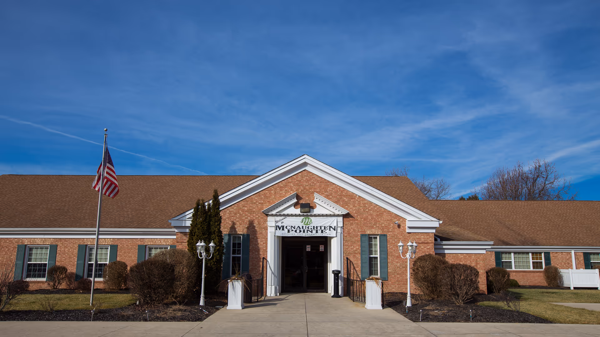 Front exterior view of McNaughten Pointe, a single-story brick building with a brown shingled roof, green window shutters, and an American flag on a flagpole to the left. The entrance has a white portico with a sign above the door reading 'McNaughten Pointe'. There are bushes and small trees in front of the building under a clear blue sky.