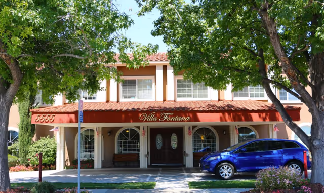 Front exterior view of Villa Fontana Retirement Community building with a red awning displaying the name, arched windows, double wooden doors, a blue car parked in front, and trees providing shade.