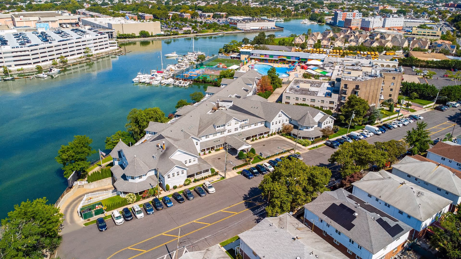 Aerial view of Sunrise at Mill Basin senior living facility located next to a body of water with boats docked nearby. The facility consists of multiple connected buildings with gray roofs, surrounded by parking lots, trees, and nearby residential and commercial buildings.