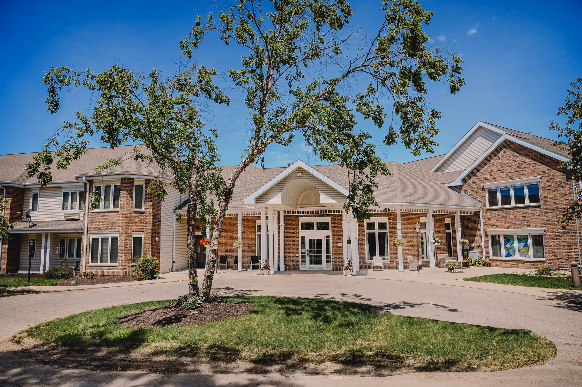 Exterior view of a senior living facility building with brick and siding walls, a covered entrance with white columns, and a tree in the foreground under a clear blue sky.